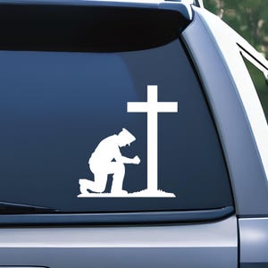 May include: White silhouette of a person kneeling in prayer in front of a cross. The person is wearing a hat and has their hands clasped together. The cross is tall and has a simple design.