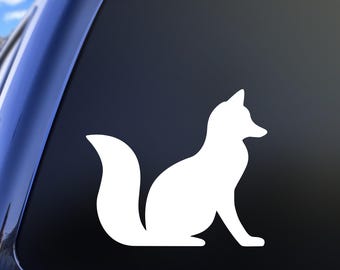 May include: A white vinyl decal featuring the silhouette of a fox sitting in profile. The fox is depicted with its ears pointed upward, a bushy tail curved behind its body, and its head turned slightly to the right, showing a clean, solid shape.