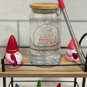 May include: A clear glass water bottle with a bamboo lid and a red silicone straw. The bottle has a Hogwarts castle graphic with a red outline. The bottle is sitting on a wooden shelf with two pink gnomes and three small Christmas trees.