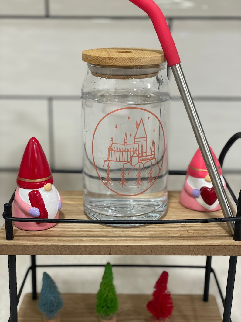 May include: A clear glass water bottle with a bamboo lid and a red silicone straw. The bottle has a Hogwarts castle graphic with a red outline. The bottle is sitting on a wooden shelf with two pink gnomes and three small Christmas trees.