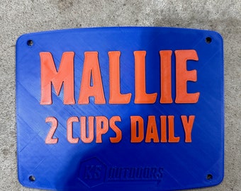 May include: Blue plastic sign with the name Mallie and the text "2 cups daily" in orange lettering. The sign has two holes for hanging and a small logo in the bottom right corner.