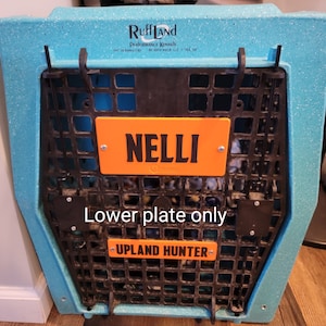 May include: A blue speckled dog crate with a black wire front. An orange plate with the name "NELLI" is attached to the front. Below, another orange plate reads "UPLAND HUNTER". The crate is made by Ruff Land Performance Kennels.