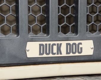 May include: A black metal dog crate with a beige plastic door. The door has a metal grill and a beige nameplate with the text "DUCK DOG" in black letters.