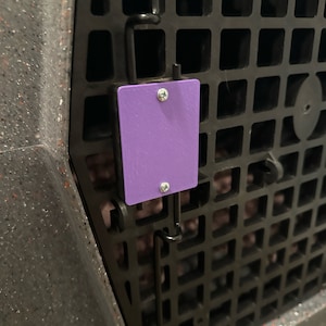 May include: Close-up of a black plastic pet carrier with a grid pattern. A small, rectangular purple tag is attached with two screws. The carrier has a speckled gray and orange exterior.