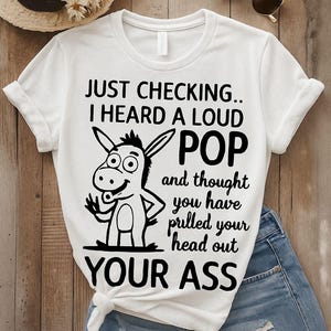 Puede incluir: Camiseta blanca con un gráfico y texto humorísticos. El diseño presenta un burro de dibujos animados y la frase "JUST CHECKING.. I HEARD A LOUD POP and thought you have pulled your head out YOUR ASS."