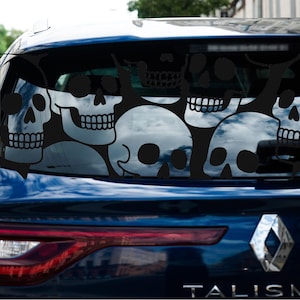 May include: Black skull window decal applied to the rear window of a blue car. The decal features a series of stylized skulls in various sizes and orientations, creating a spooky and eye-catching design. The car's taillight is visible.