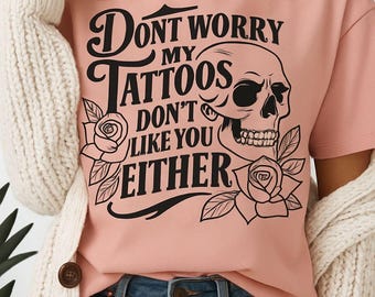 May include: A light pink t-shirt with the text "Don't worry my tattoos don't like you either" in black, along with a skull and rose graphic. The shirt is paired with blue jeans.