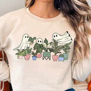 May include: A cream-colored sweatshirt with a graphic of ghosts holding potted plants. The ghosts are white and the plants are green with brown pots.