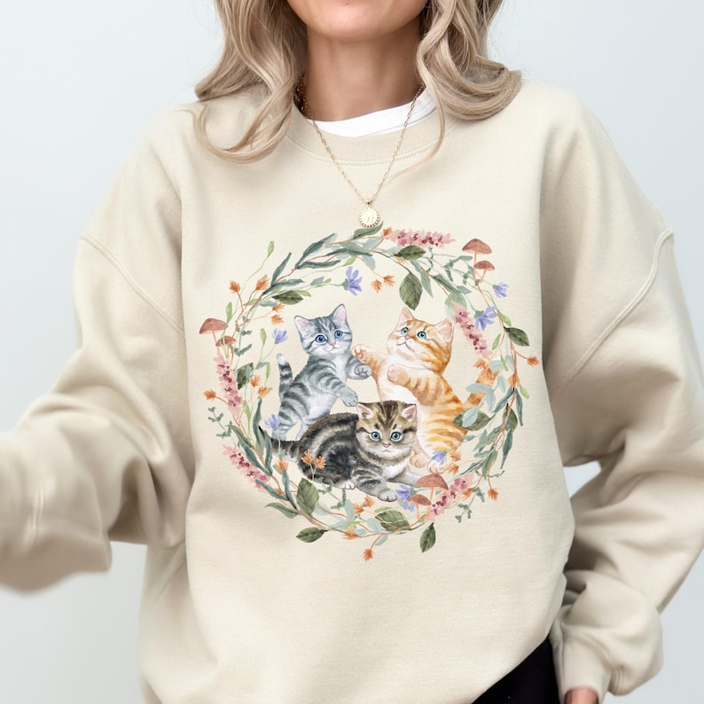 May include: A beige sweatshirt featuring a design of three kittens playing in a wreath of flowers. The kittens are grey, orange, and tabby.