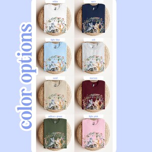 May include: A range of colour options for a sweatshirt featuring a graphic of three cats in a floral wreath. The colours include white, navy, light blue, ash, sand, maroon, military green, and light pink.