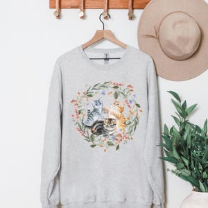 May include: A grey sweatshirt with a floral wreath design featuring three kittens. The kittens are grey, brown, and orange. The wreath is made of green leaves, pink and white flowers, and brown mushrooms.