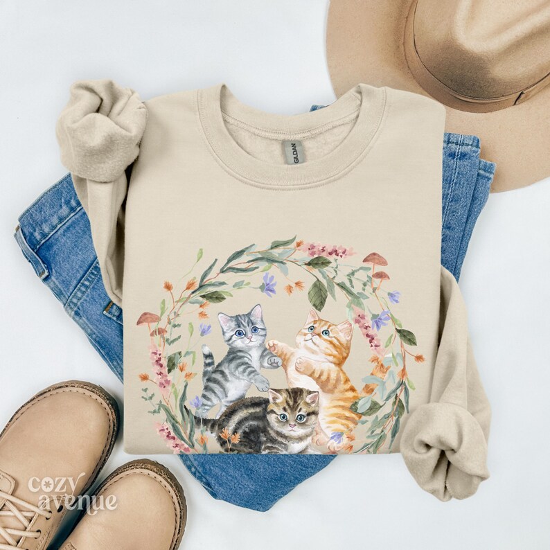 May include: A beige sweatshirt featuring a floral wreath design with three kittens playing. The kittens are grey, orange, and tabby. The wreath is made of green leaves and pink, purple, and white flowers.