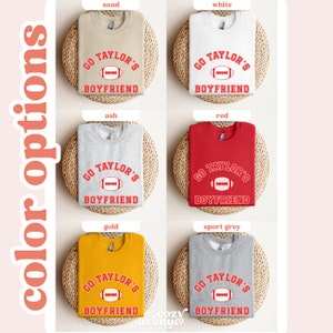 May include: A set of six sweatshirts in different colors, each featuring a red graphic of a football with the text "GO TAYLOR'S BOYFRIEND". The sweatshirts are displayed on a woven surface.