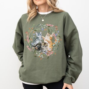 May include: A green sweatshirt with a floral wreath design featuring four kittens playing. The kittens are grey, white, orange, and brown.
