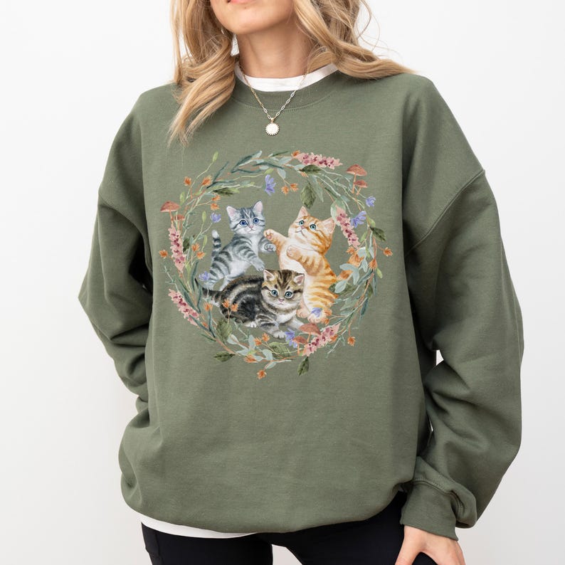 May include: A green sweatshirt with a floral wreath design featuring four kittens playing. The kittens are grey, white, orange, and brown.