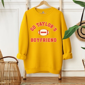 May include: Yellow sweatshirt with red text that says "Go Taylor's Boyfriend" with a red and white football graphic.