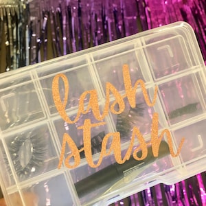 May include: A clear plastic storage box with multiple compartments labelled "lash stash" in gold glitter lettering. The box contains false eyelashes and a black mascara wand.