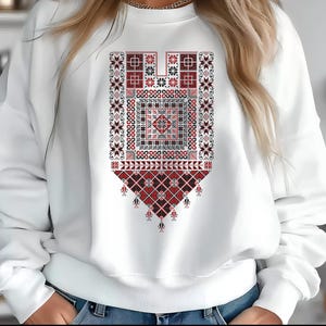 Pode incluir: Camisola branca com um design geométrico detalhado em vermelho, preto e branco. O design é centrado no peito e inclui várias formas e padrões, criando um visual tradicional bordado. A camisola tem mangas compridas.