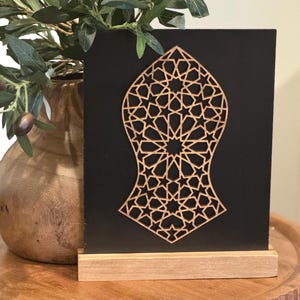May include: A decorative wooden art piece featuring a geometric star pattern, mounted on a black background and a natural wood base. The artwork is displayed on a wooden table, with a vase of olive branches in the background.