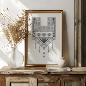 Könnte beinhalten: Ein gerahmter Druck mit einem schwarz-weißen geometrischen Muster, das an traditionelle Stickereien erinnert. Das Kunstwerk befindet sich in einem goldenen Rahmen und steht auf einem Holzschrank. Eine klare Glasvase mit getrockneten Pflanzen und eine kleine weiße Vase sind ebenfalls zu sehen.