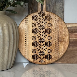May include: Round wooden cutting board with a decorative floral pattern. The board has a handle and a dark brown floral design etched into the light brown wood. A vase with greenery is in the background.