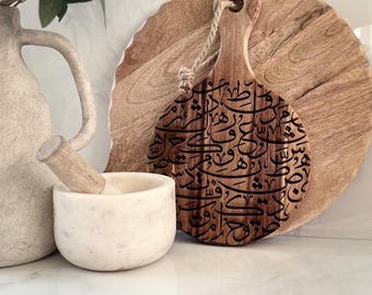 May include: A wooden serving board with a handle and a decorative rope. The board has a dark brown engraved design. A round wooden board and a white marble mortar and pestle are also in the image.