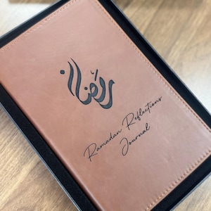 May include: A brown leather journal with the words "Ramadan Reflections Journal" printed on the cover. The journal is in a black box. The cover has an Arabic calligraphy design.