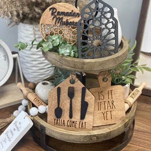 May include: A two-tiered wooden tray with decorative items. The top tier features a wooden disc with "Ramadan Mubarak" and a black lantern. The bottom tier has wooden cutouts with utensils and the text "Yalla Come Eat" and "Is it Iftar Yet".