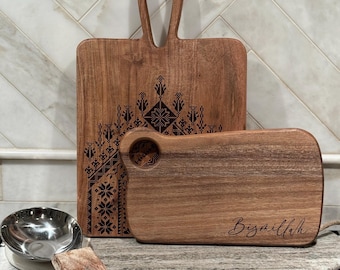 May include: Two wooden cutting boards. The larger board has a handle and a decorative black pattern. The smaller board has a round hole and the word "Bismillah" in script. A small metal bowl and a wooden spoon rest nearby.