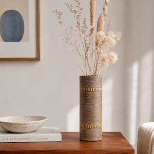 Könnte beinhalten: Eine hohe, zylindrische Keramikvase mit einer texturierten, dunkelbraunen Oberfläche und horizontalen, gerippten Bändern in einem gedämpften Goldton. Die Vase ist mit einer Sammlung getrockneter Gräser und beiger botanischer Stängel gefüllt.