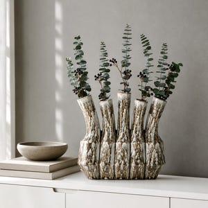 Könnte beinhalten: Eine mehrhalsige Keramikvase mit einer texturierten, gesprenkelten braun-weißen Glasur. Die Vase hat fünf einzelne vertikale Öffnungen, in denen jeweils ein Zweig grüner Eukalyptus mit kleinen dunklen Beeren steckt, was ein strukturiertes Arrangement ergibt.