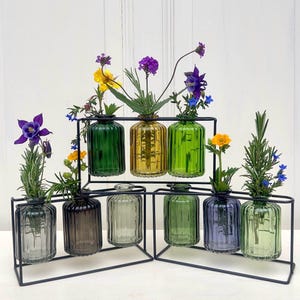 May include: A set of nine colorful glass bud vases arranged in a black metal frame. Each vase holds a different flower, showcasing a variety of colors including purple, yellow, green, and blue. The vases are ribbed and have a vintage aesthetic.