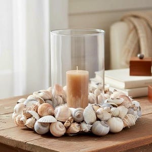 Könnte beinhalten: Ein zylindrischer Kerzenhalter aus klarem Glas steht in der Mitte eines kreisförmigen Kranzes aus verschiedenen natürlichen Muscheln. Eine einzelne beige Stumpenkerze befindet sich im Glashalter, umgeben von Muscheln in Weiß-, Beige- und Grautönen.