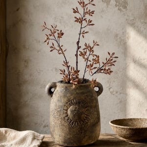 Könnte beinhalten: Eine texturierte, dunkelbraune Keramikvase mit zwei kleinen seitlichen Griffen und einem erhabenen Sonnenemblem auf der Vorderseite. Die Vase enthält einen Zweig mit kleinen, getrockneten, rotbraunen Blättern. Die Oberfläche hat ein mattes Finish.