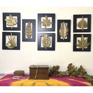 May include: A set of eight gold leaf wall decor pieces in black frames. The leaves are in various shapes and sizes, and they are arranged in a symmetrical pattern on a white wall.