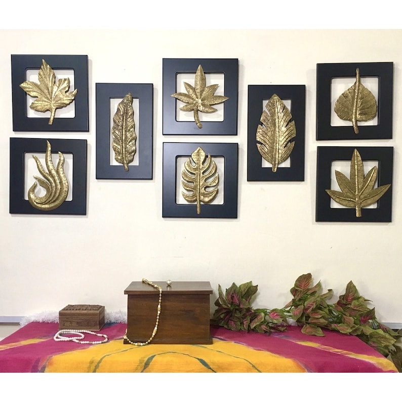 May include: A set of eight gold leaf wall decor pieces in black frames. The leaves are in various shapes and sizes, and they are arranged in a symmetrical pattern on a white wall.
