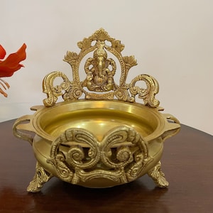 May include: A gold metal incense burner with intricate details and a depiction of the Hindu deity Ganesha on the top. The burner has three legs and a round bowl.