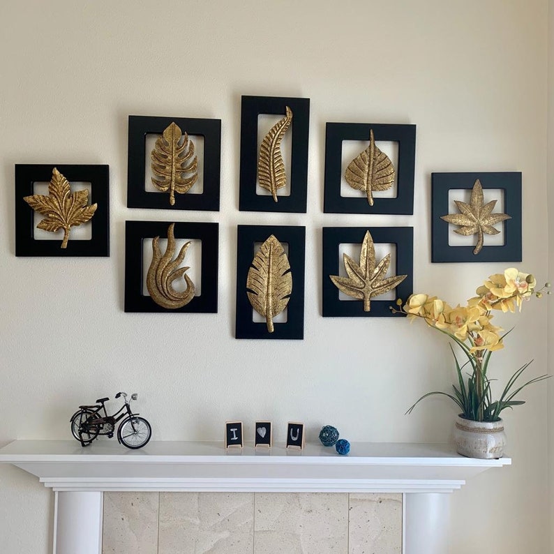 May include: A wall decor set of eight black framed gold leaf wall art pieces featuring various leaf designs. The frames are arranged in a grid pattern on a white wall.