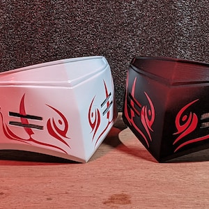 May include: Two masks, one white and one black, with red tribal designs. The masks have a strap on the back and a raised design on the front.