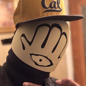 May include: A mustard yellow baseball cap with the word Cal embroidered in navy blue, worn over a beige face mask featuring a black hand-shaped cutout design with a central eye graphic, paired with a black neck gaiter and a black quilted jacket.