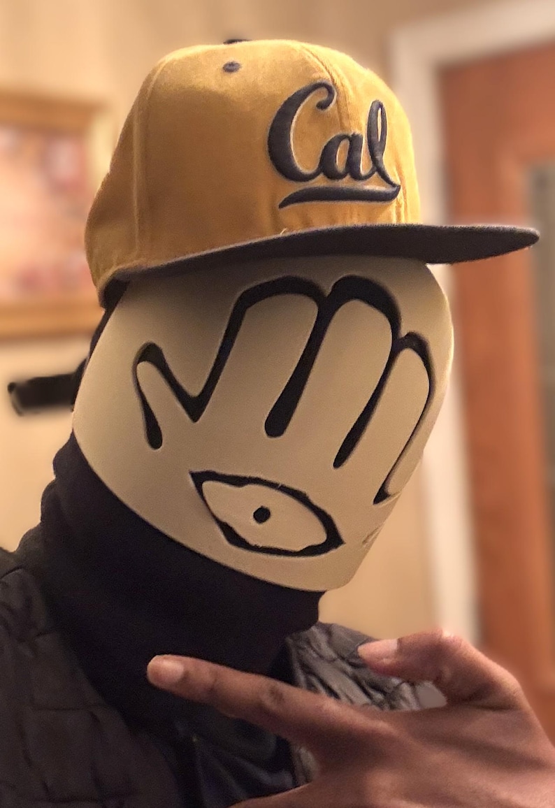 May include: A mustard yellow baseball cap with the word Cal embroidered in navy blue, worn over a beige face mask featuring a black hand-shaped cutout design with a central eye graphic, paired with a black neck gaiter and a black quilted jacket.