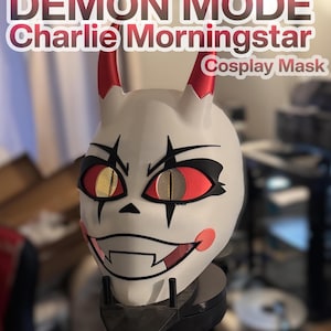 Pode incluir: Uma máscara de cosplay branca com chifres vermelhos, olhos vermelhos e amarelos e um sorriso sinistro. A máscara tem detalhes pretos ao redor dos olhos e da boca. O texto na máscara diz "DEMON MODE Charlie Morningstar Cosplay Mask."