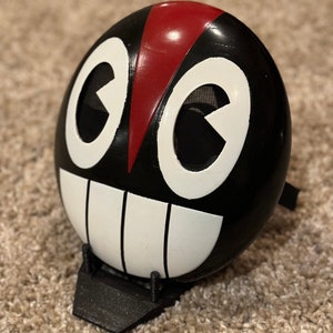 Può includere: Una maschera nera e bianca con una striscia rossa al centro. La maschera ha due grandi occhi bianchi con una rete nera dietro e un ampio sorriso bianco con linee nere.