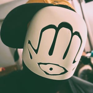 May include: A mustard yellow baseball cap worn with a white face mask featuring a cutout design of a hand with an eye symbol in the palm. The mask is smooth and covers the front of the face, while the cap has a dark brim and a structured crown.