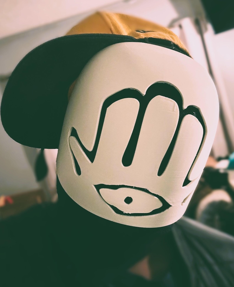 May include: A mustard yellow baseball cap worn with a white face mask featuring a cutout design of a hand with an eye symbol in the palm. The mask is smooth and covers the front of the face, while the cap has a dark brim and a structured crown.