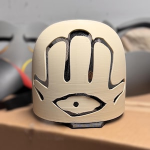 May include: A cream-coloured, dome-shaped mask featuring a cutout design of a hand with an eye in the centre. The cutout areas are backed with a black mesh material, providing a contrast to the smooth, light-coloured surface of the mask structure.