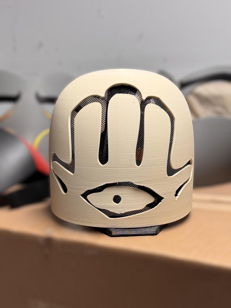 May include: A cream-coloured, dome-shaped mask featuring a cutout design of a hand with an eye in the centre. The cutout areas are backed with a black mesh material, providing a contrast to the smooth, light-coloured surface of the mask structure.