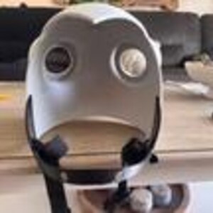 May include: A white, rounded protective helmet featuring two circular eye openings with dark, recessed lenses. The helmet has a smooth, curved exterior shell and includes a black adjustable chin strap attached to the lower sides for secure fastening.