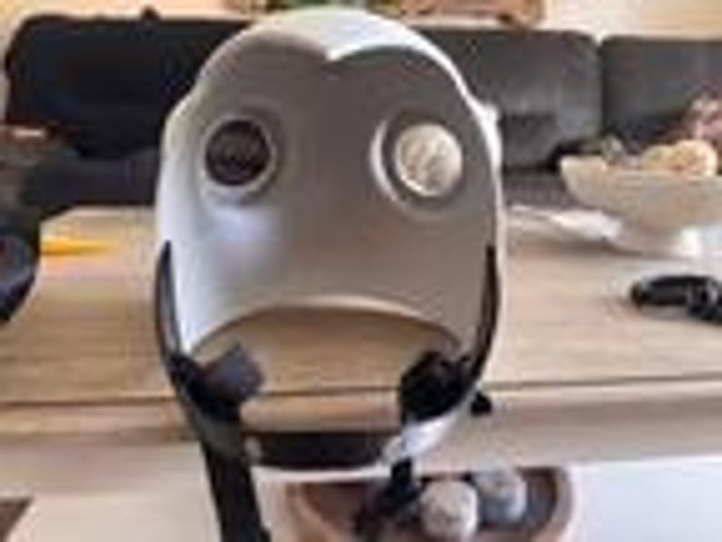 May include: A white, rounded protective helmet featuring two circular eye openings with dark, recessed lenses. The helmet has a smooth, curved exterior shell and includes a black adjustable chin strap attached to the lower sides for secure fastening.