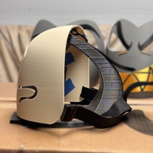 May include: A beige 3D printed mask with a smooth exterior and a black interior frame. The mask features a black adjustable elastic strap attached to the sides and small pieces of blue painter's tape visible on the inner structure of the mask.